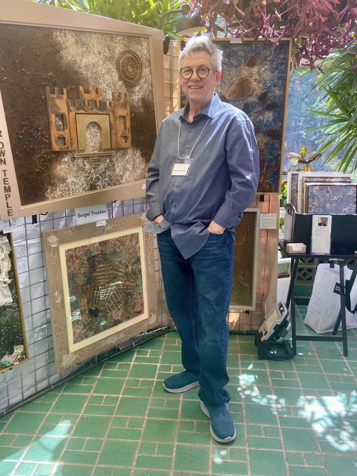 Sergei Troshin at an art fair, standing in front of his mixed media paintings